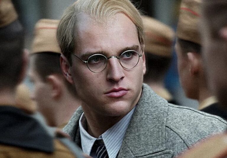 Jonas Dassler como Dietrich Bonhoeffer em "A Redenção: A História Real de Bonhoeffer" (2024). Na imagem, o ator está caracterizado como o teólogo alemão, com cabelo loiro, óculos redondos e expressão séria, trajando um casaco cinza sobre uma camisa e gravata. Ele aparece cercado por homens uniformizados, sugerindo um cenário da Alemanha nazista durante a Segunda Guerra Mundial.