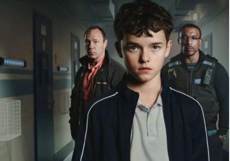 A imagem mostra um garoto jovem de expressão séria e determinada no centro, vestindo uma jaqueta esportiva azul escura com detalhes brancos e uma camisa polo cinza. Atrás dele, há dois homens adultos, um aparentando ser um policial com um colete tático e outro com uma aparência mais casual, usando uma jaqueta e camisa vermelha. O cenário parece ser um corredor de um centro de detenção ou prisão juvenil, com portas metálicas e iluminação fria. A composição da imagem sugere um drama policial ou uma narrativa intensa envolvendo o garoto e as figuras de autoridade ao fundo.