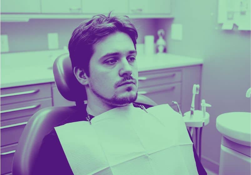A imagem mostra um homem sentado em uma cadeira de dentista, vestindo o avental descartável típico de procedimentos odontológicos. Ele tem expressão séria e parece exausto ou introspectivo, olhando fixamente para a frente, sem sorrir. O ambiente ao redor é uma sala de atendimento odontológico, com móveis planejados, equipamentos clínicos e iluminação suave. A imagem possui um filtro em tons de verde e roxo, dando-lhe um ar estilizado e reflexivo. Essa combinação de elementos transmite uma sensação de desgaste emocional e resistência silenciosa — muito alinhada ao relato de um autista lidando com desafios na saúde bucal.