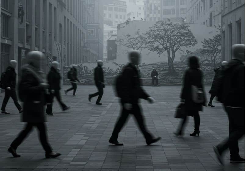 A imagem mostra uma cena urbana em tons de cinza, transmitindo uma atmosfera fria e mecânica. Várias pessoas caminham apressadas em diferentes direções, com rostos desfocados ou apagados, o que simboliza a perda de identidade e individualidade. Todas parecem alheias umas às outras, como engrenagens em movimento automático. Ao fundo, há um grande mural com a ilustração de uma paisagem natural, com árvores — um forte contraste com o concreto e a pressa ao redor. Diante dele, uma única figura está sentada em silêncio, imóvel, em postura contemplativa, sugerindo um momento de pausa e reflexão em meio ao caos produtivista. A imagem evoca exatamente o conflito entre o "fazer" frenético da vida moderna e a necessidade esquecida de simplesmente "ser".