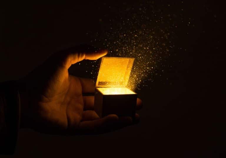 A imagem mostra uma mão segurando uma pequena caixa aberta, de onde escapa uma luz dourada intensa, que se espalha em partículas brilhantes pelo ar. O fundo é completamente escuro, destacando o contraste entre a luz e as sombras, criando uma atmosfera de mistério, reverência e beleza. A cena remete à ideia de algo precioso, imensurável e que não pode ser contido.