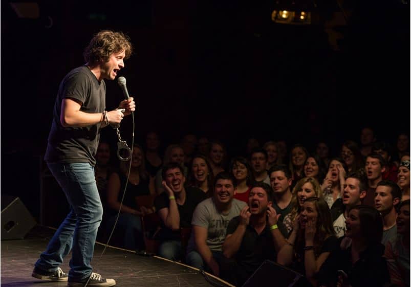A imagem mostra um comediante se apresentando em um palco de stand-up comedy. Ele segura um microfone com uma das mãos enquanto a outra está algemada, com uma algema solta pendendo do pulso — um símbolo visual forte, sugerindo a ideia de censura, prisão ou limites à liberdade de expressão. O comediante veste roupas casuais: camiseta preta, jeans e tênis. Ele aparenta estar envolvido emocionalmente com a performance, talvez fazendo uma piada intensa ou provocadora. A plateia à sua frente está lotada e demonstra reações diversas: algumas pessoas estão rindo entusiasticamente, outras parecem surpresas ou desconfortáveis. Essa diversidade de expressões reforça a ambiguidade do humor apresentado e a polarização que ele pode causar. A iluminação foca no comediante, deixando o fundo mais escuro, o que destaca ainda mais a figura central e cria uma atmosfera de tensão dramática misturada ao entretenimento. A cena representa visualmente o debate sobre os limites do humor, os riscos da transgressão em apresentações cômicas e o impacto do discurso cômico no público.
